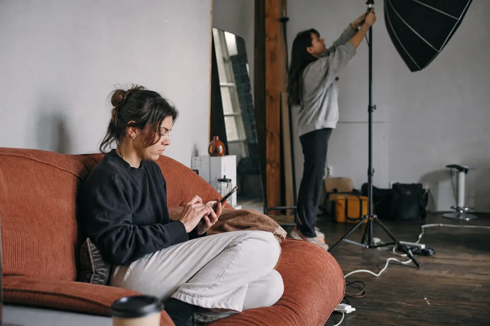 Two adult creators in a small studio, one signing a consent on her phone while the other sets up a light