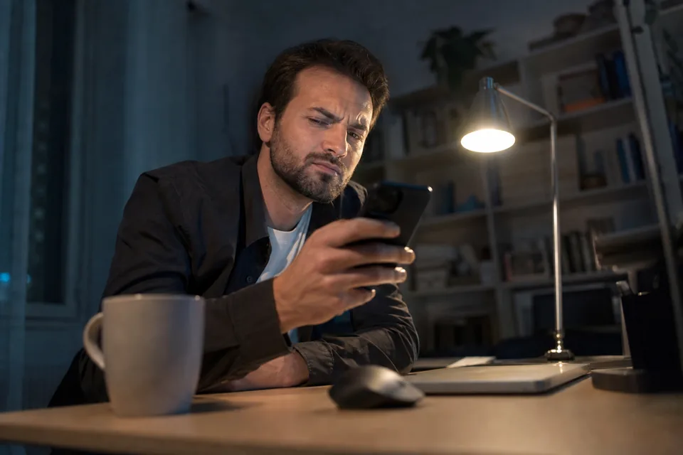 Man at a desk looking at his phone with a skeptical frown